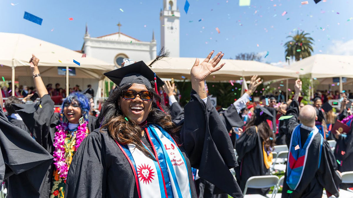 A graduate waving to the camera as other graduates cheer and confetti falls around them